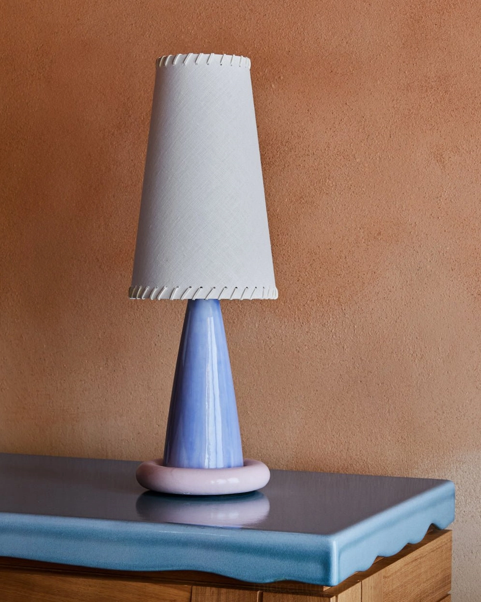 Modern ribbon table lamp displayed on terracotta accent tabletop with light grey shade, blue ceramic base, and pink ring foot