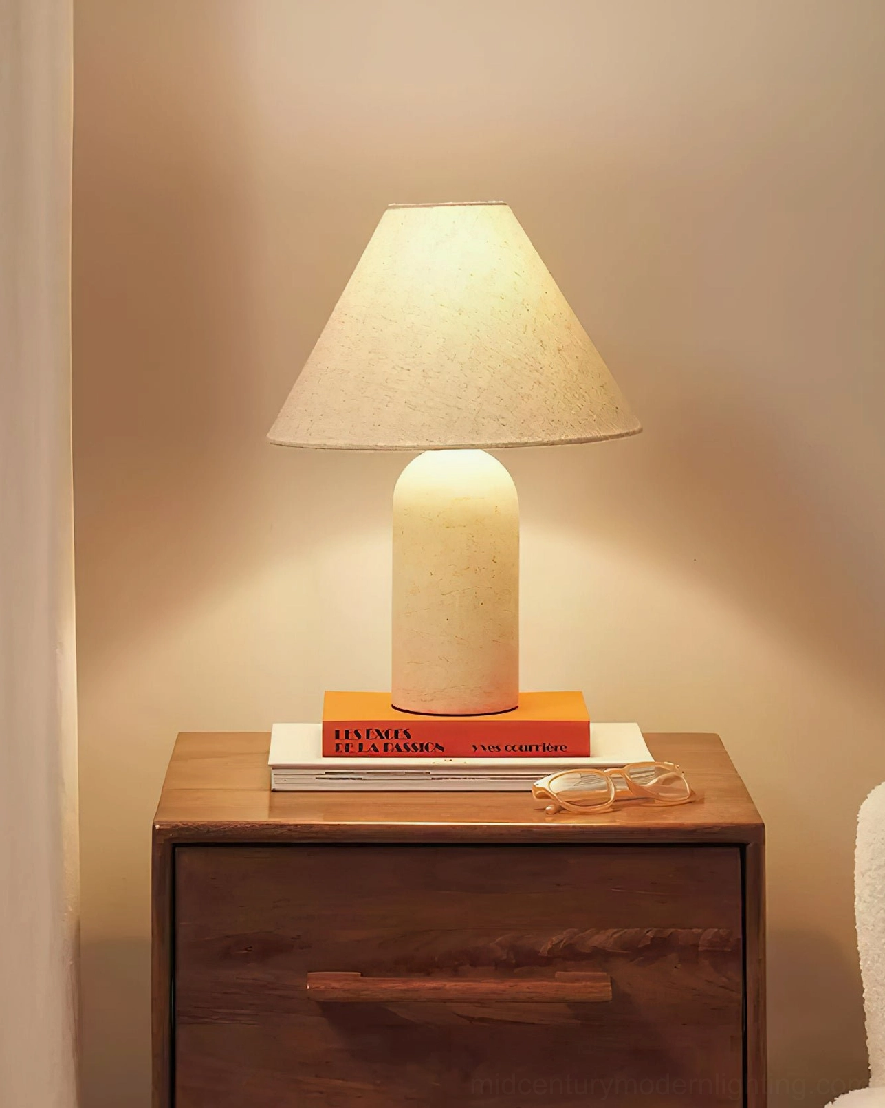 Off-white cylindrical marble lamp with beige fabric shade on warm-toned bedside setup, stacked books and eyeglasses, intimate bedroom lighting