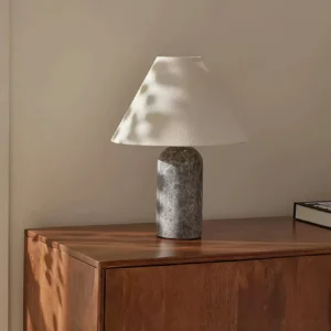 Grey marble table lamp with natural veining on sunny bedroom dresser, warm ambient lighting, three-quarter side view showing conical linen shade