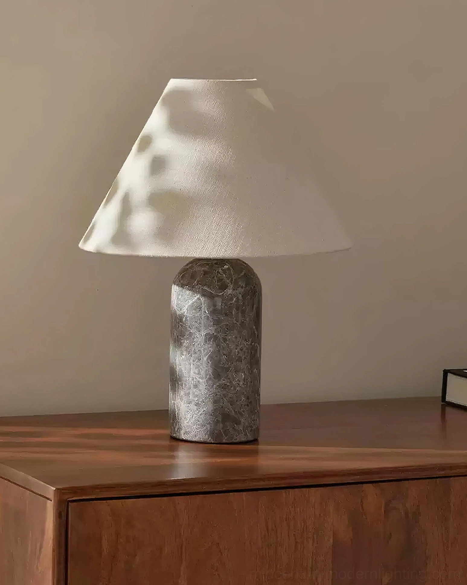 Grey speckled marble table lamp base with beige conical fabric shade on sunlit wooden dresser corner, three-quarter side view
