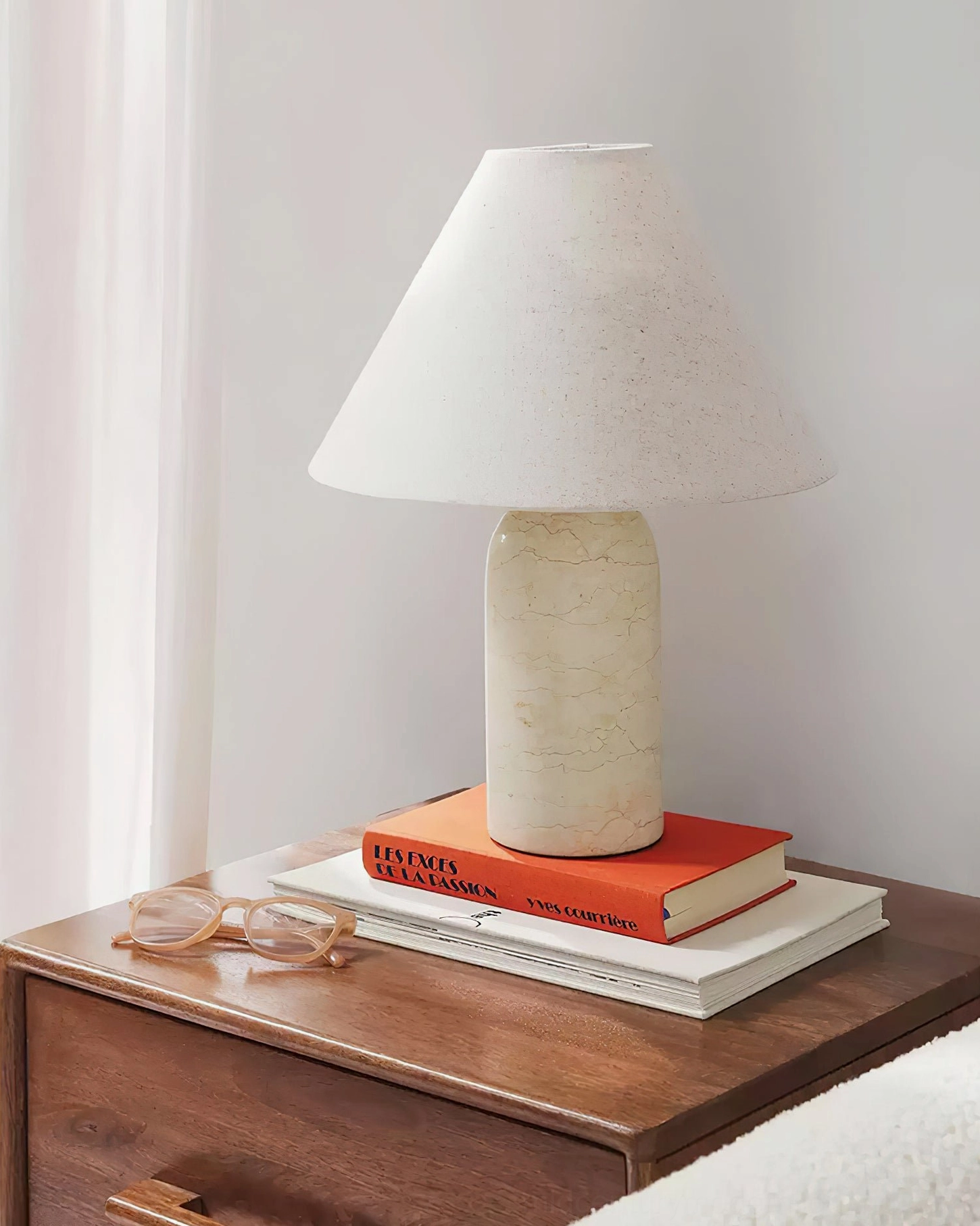 Off-white cylindrical marble base lamp with white conical fabric shade on bedroom bedside table, stacked books and eyewear, bedside lighting