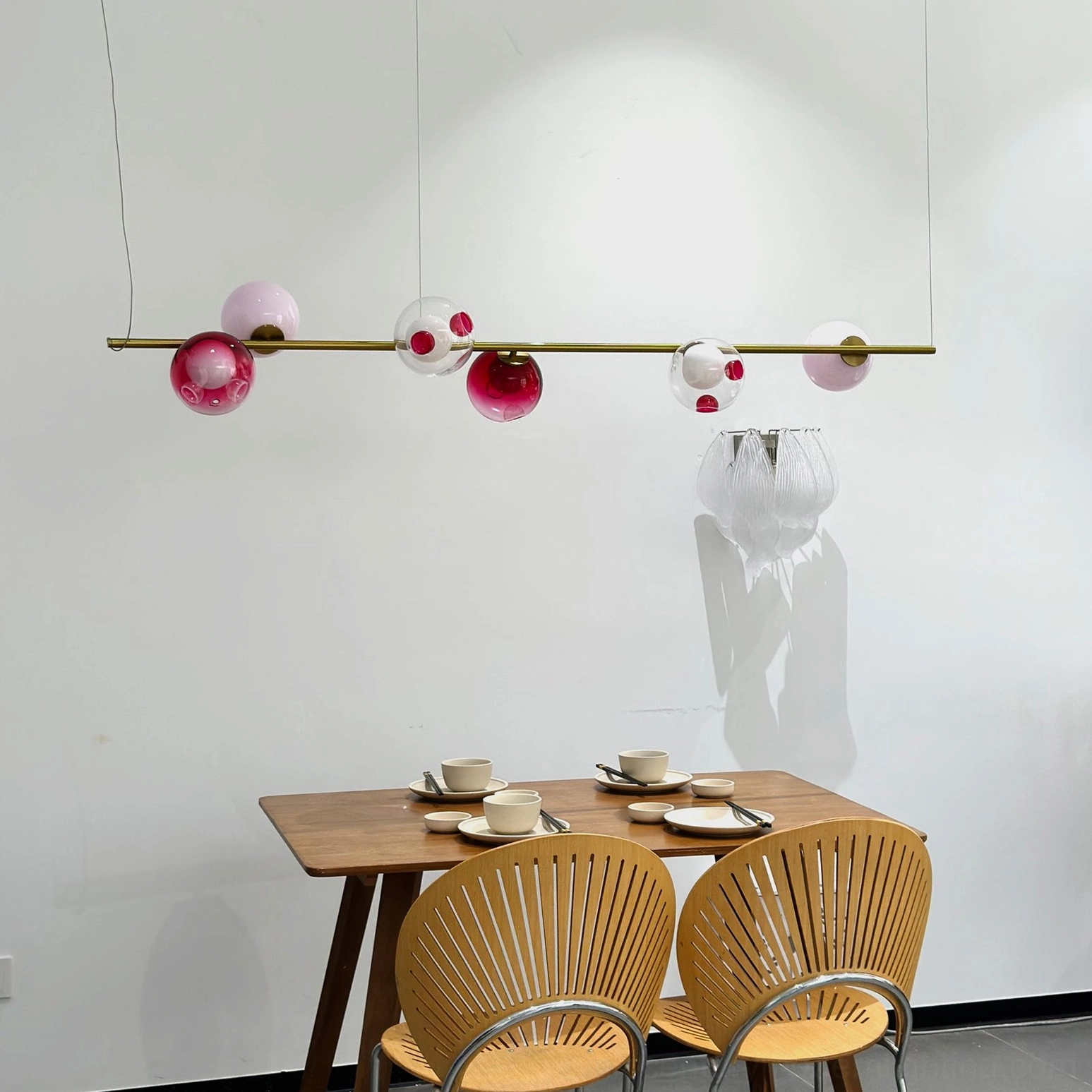 Sedna linear pendant light installed above dining table with wooden chairs and rattan seating - overhead view showing red and clear glass details