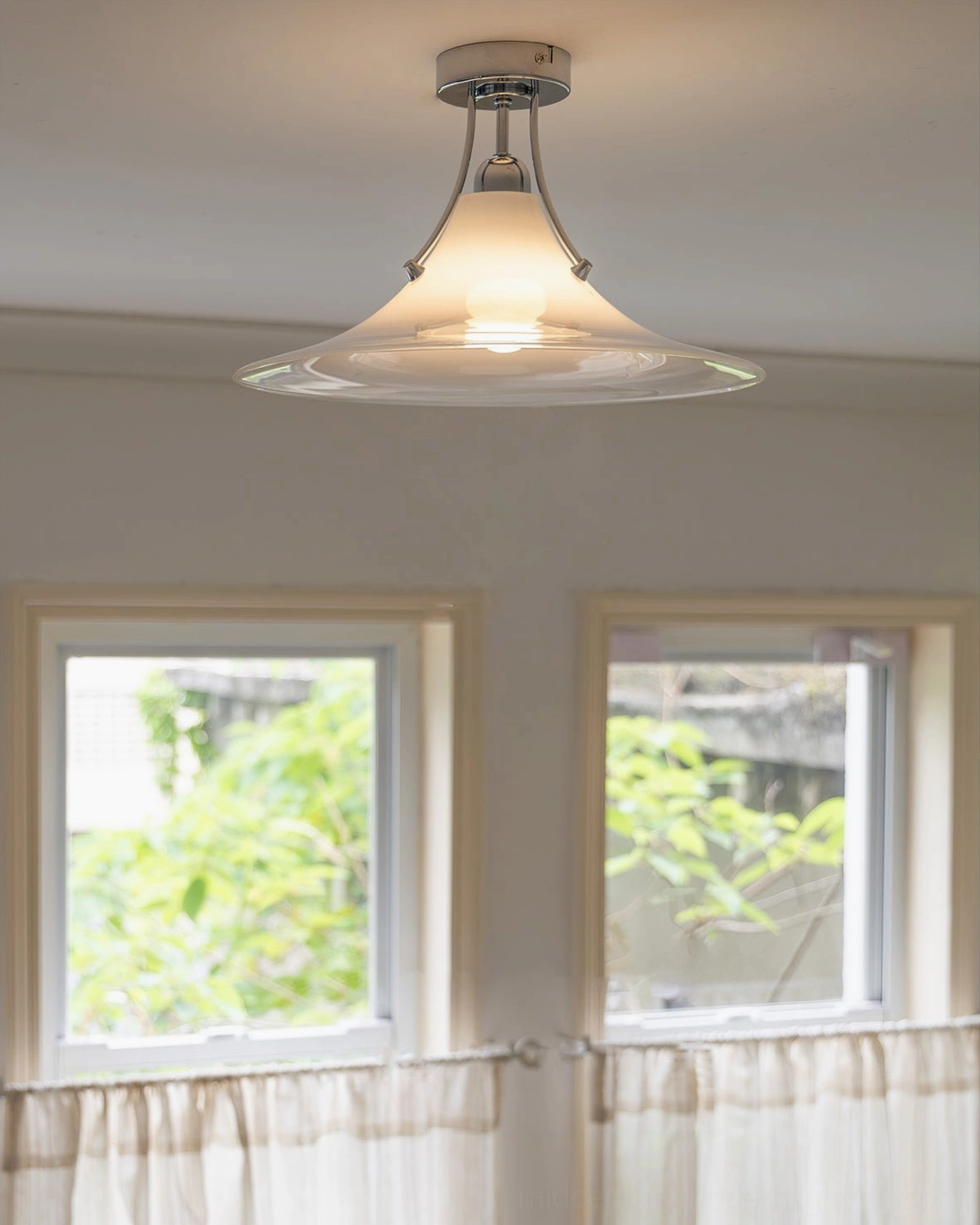 Modern ceiling lamp installed in sunlit room with double windows and sheer curtains neutral home decor