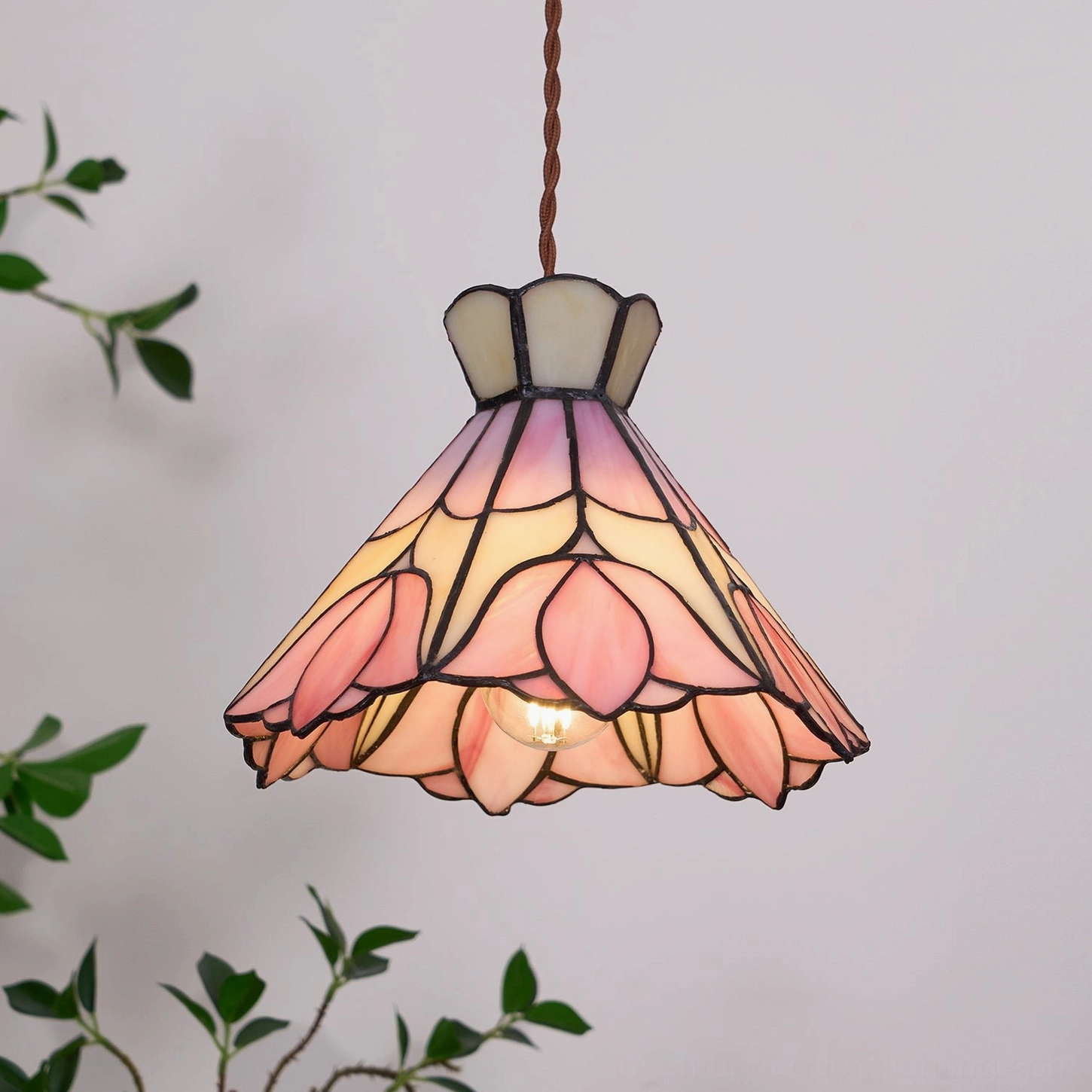 Lit Serena stained glass pendant lamp displaying exposed bulb with green plant backdrop in home setting