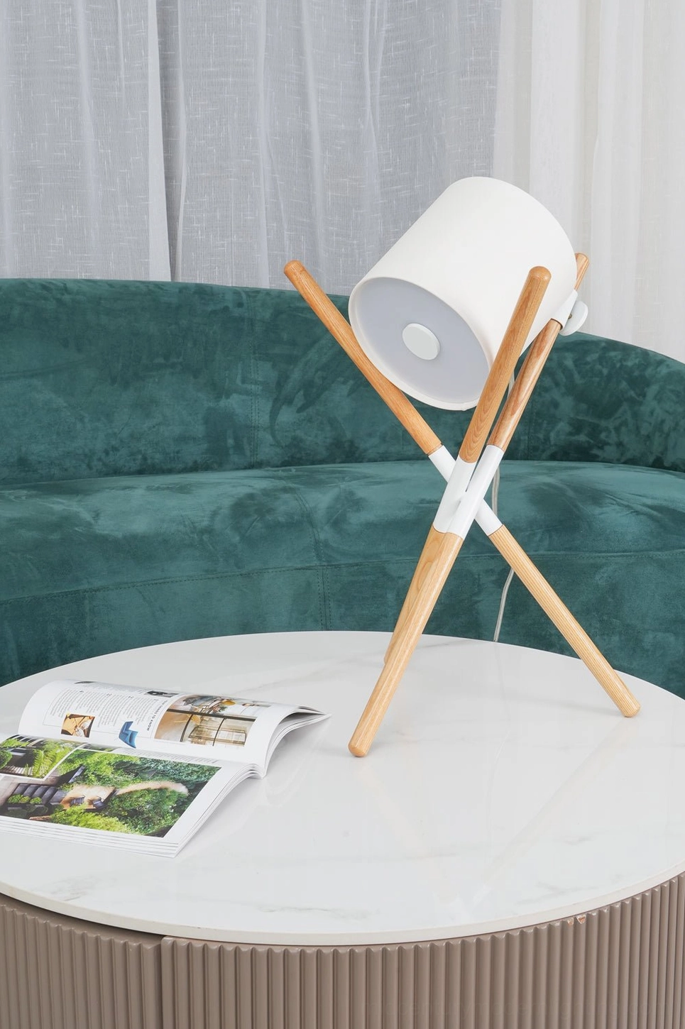 Light wood tripod table lamp with white shade on white round side table in modern living room with green velvet sofa