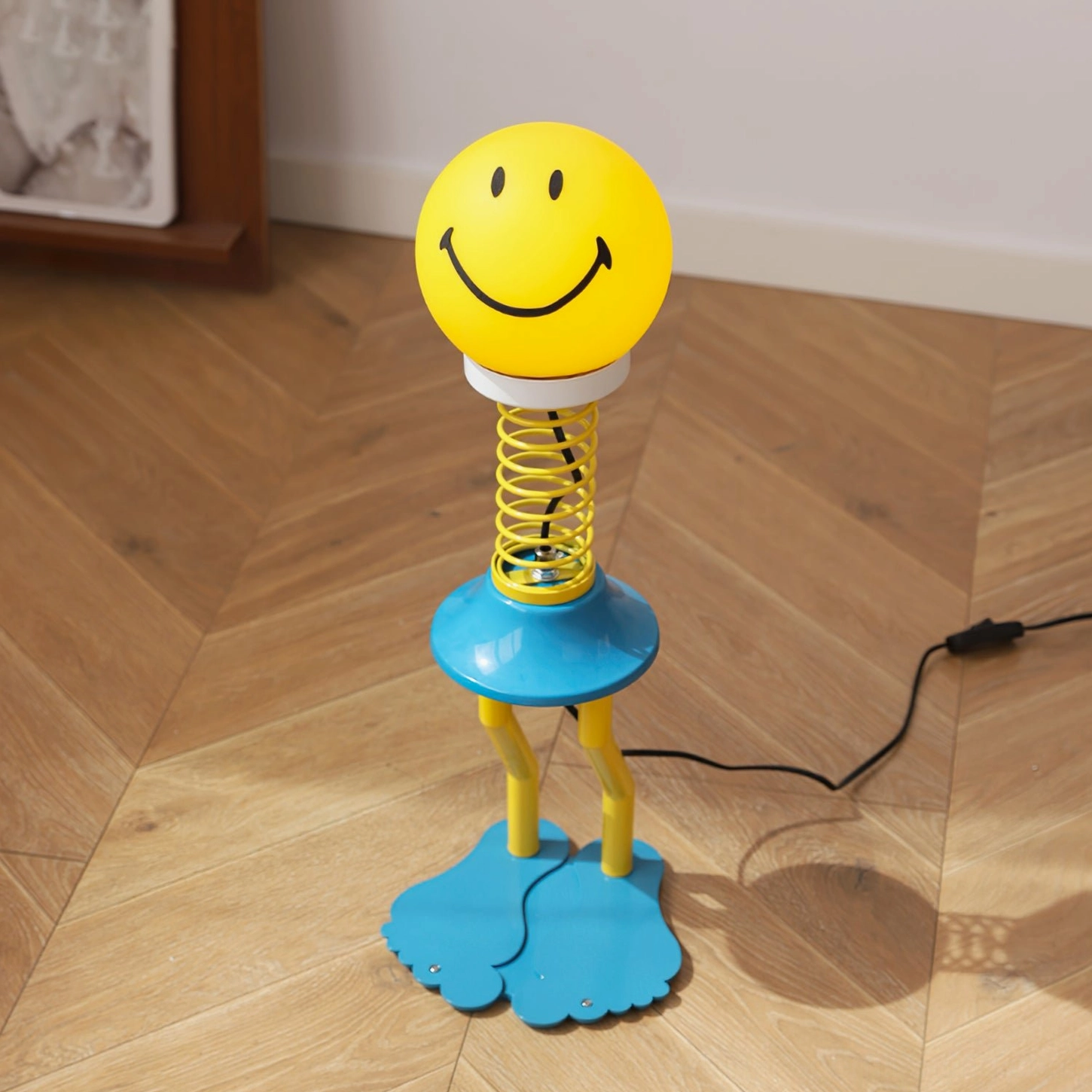 Low angle side view of spiral spring smiley lamp showing metal stem and blue dual feet on herringbone flooring