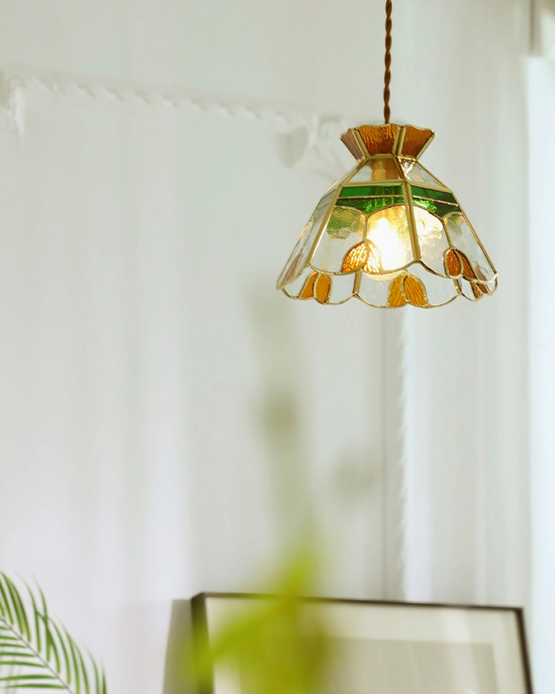 Side view of handcrafted vitral pendant lamp with sheer curtain backdrop and green plant foreground