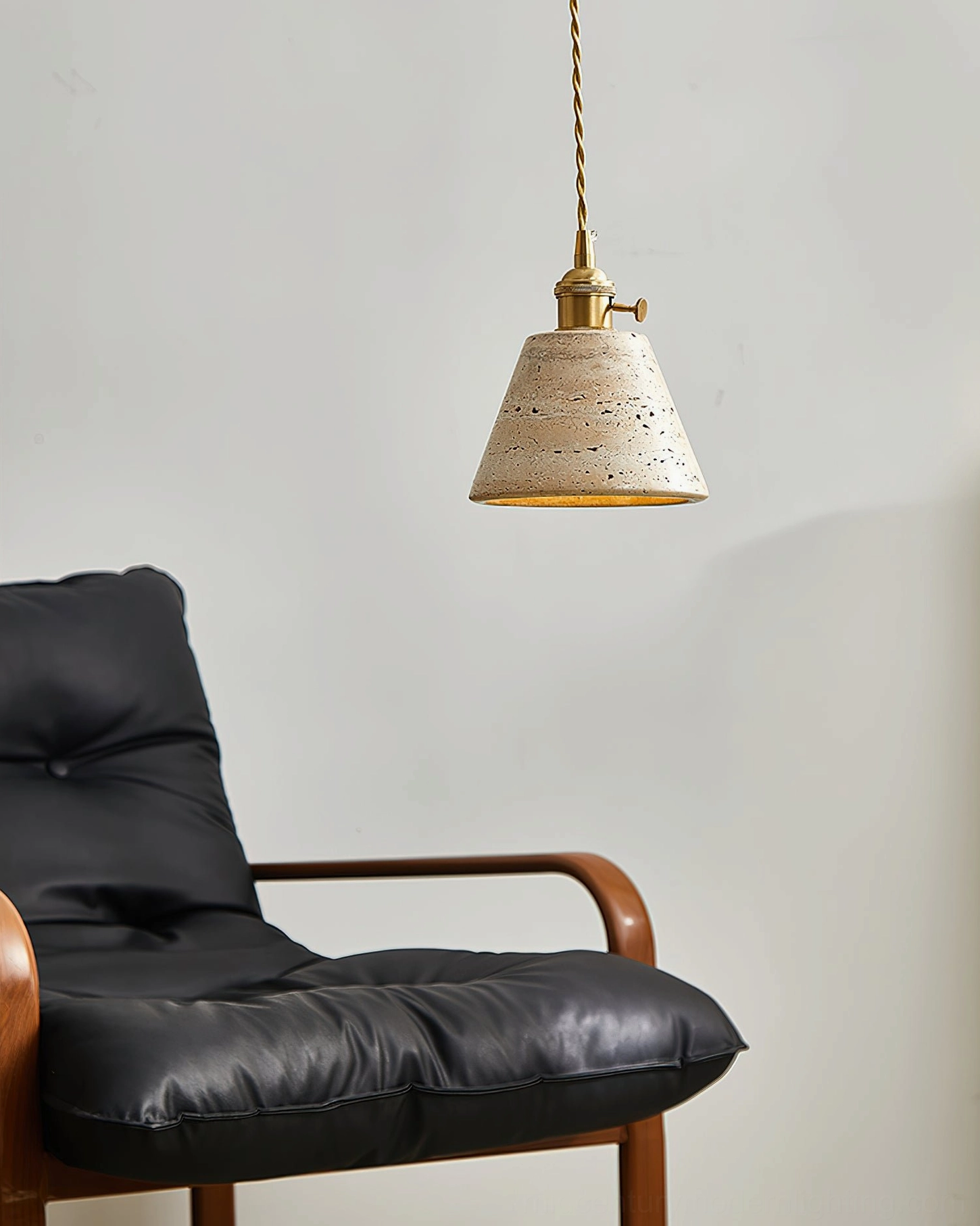 Stella conical travertine pendant light in minimalist living room beside black leather lounge chair, showcasing natural porous stone texture and brass accents