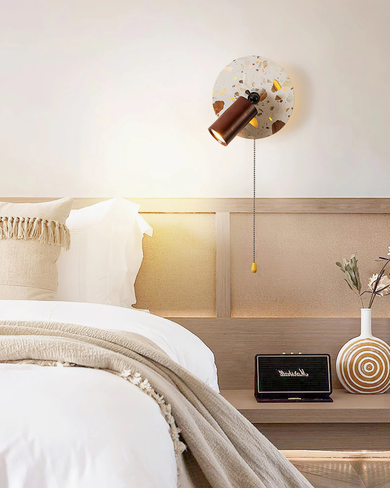 Swizzle wall lamp in master bedroom above bedside shelf with warm illumination complementing wooden headboard and soft furnishings