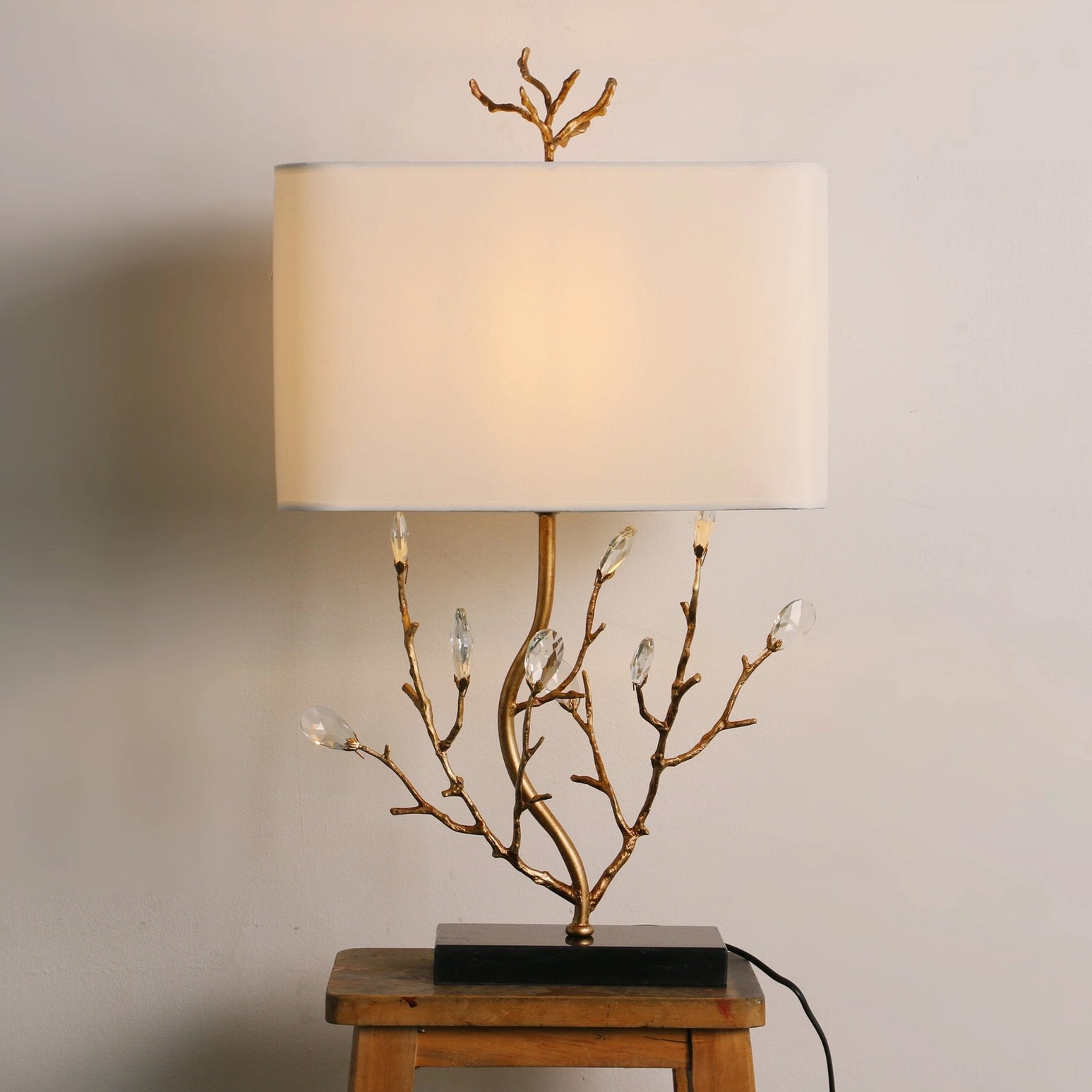 Lit Sylvia branch table lamp creating warm ambient glow, illuminated fabric shade with black square base on wooden stool