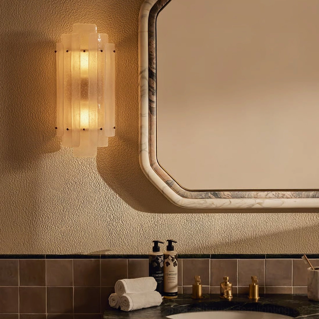 Tasha wall sconce illuminated in bathroom vanity setting with marble mirror frame and warm glow
