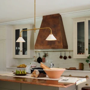 Theron brass pendant chandelier installed over kitchen island in farmhouse kitchen, showing warm brass hardware and off-white cone shades with natural lighting