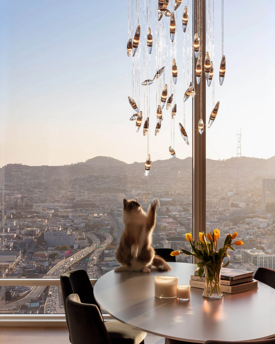 Tiered metal droplet chandelier above round wooden table with city mountain view on top-floor rooftop dining area