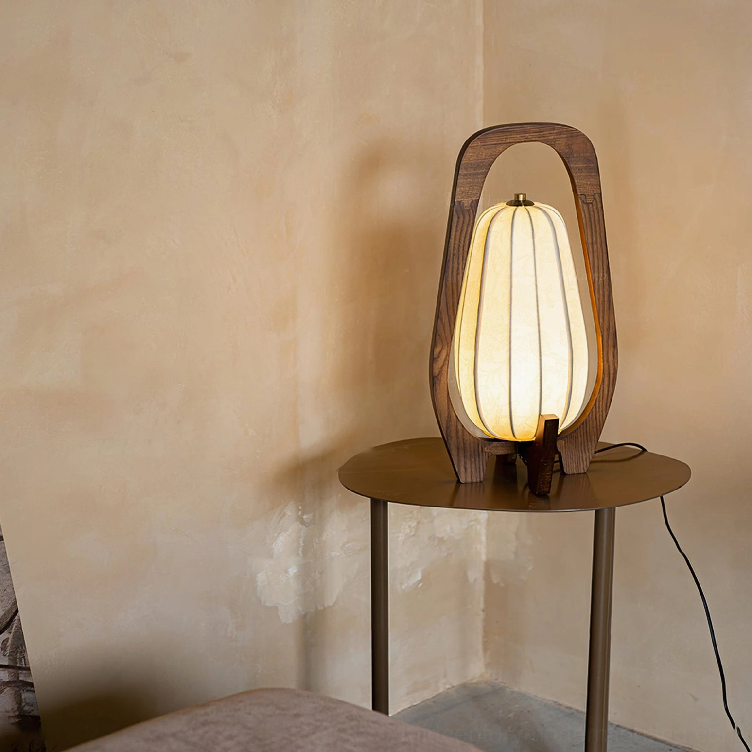 Totem wood table lamp on round side table against textured wall, side view with visible power cord and brass fixture