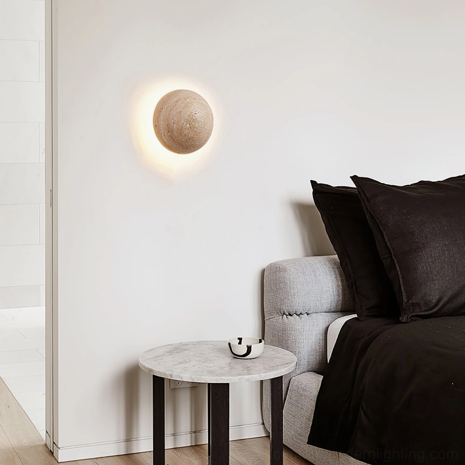 Round travertine wall lamp with glowing halo effect in bedroom with grey sofa and black bedding