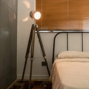 Mid-century modern tripod floor lamp positioned in vintage bedroom with iron bed, warm LED lighting casting shadows on weathered wood tripod legs