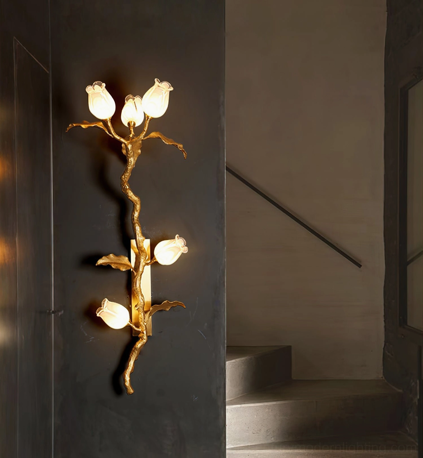 Tulip Branch Brass Wall Sconce installed in stairwell with warm glowing lights and dark accent wall creating sculptural presence