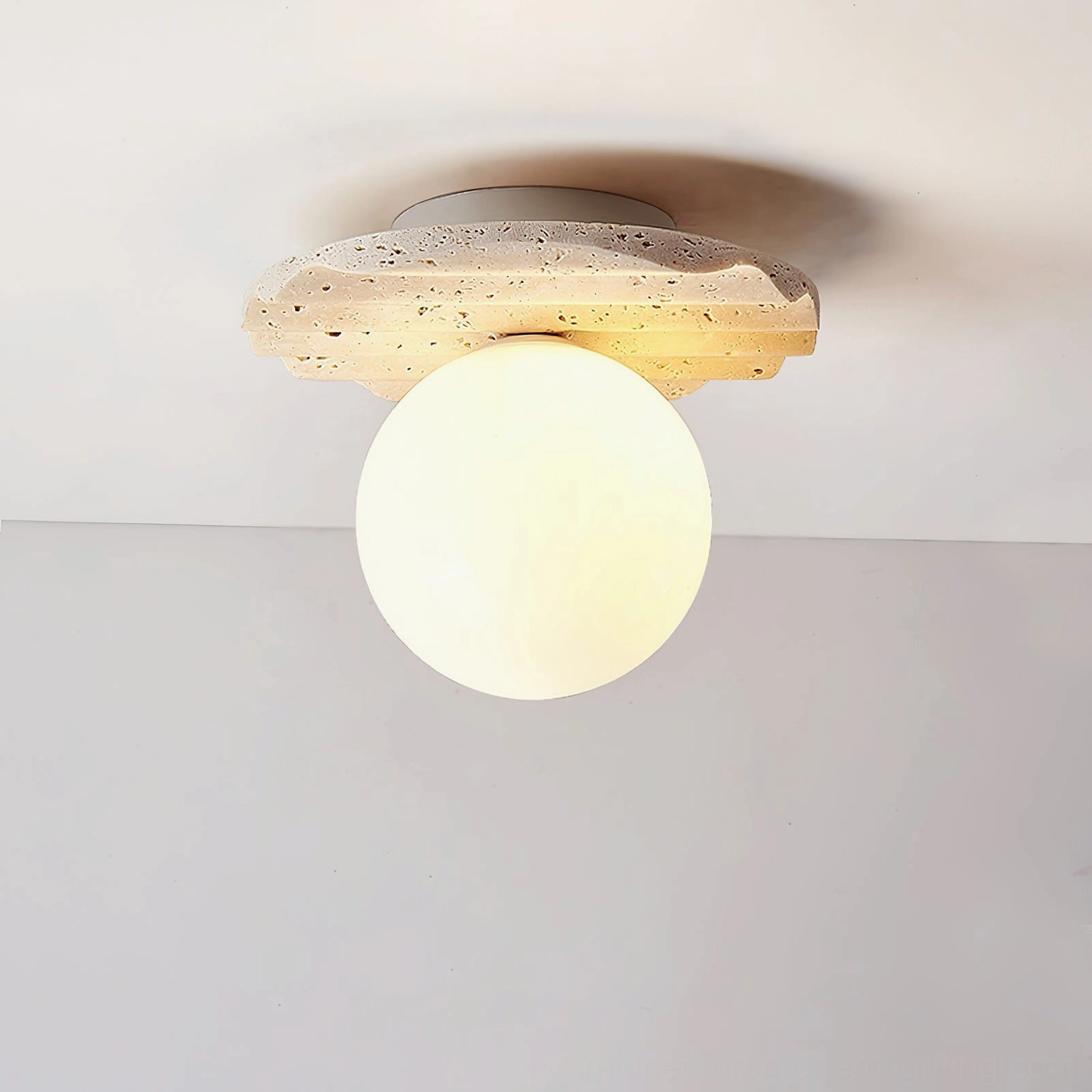 Victor travertine light fixture from side angle, illuminated, showing scalloped stone edge profile and warm light cast from glass globe, architectural perspective