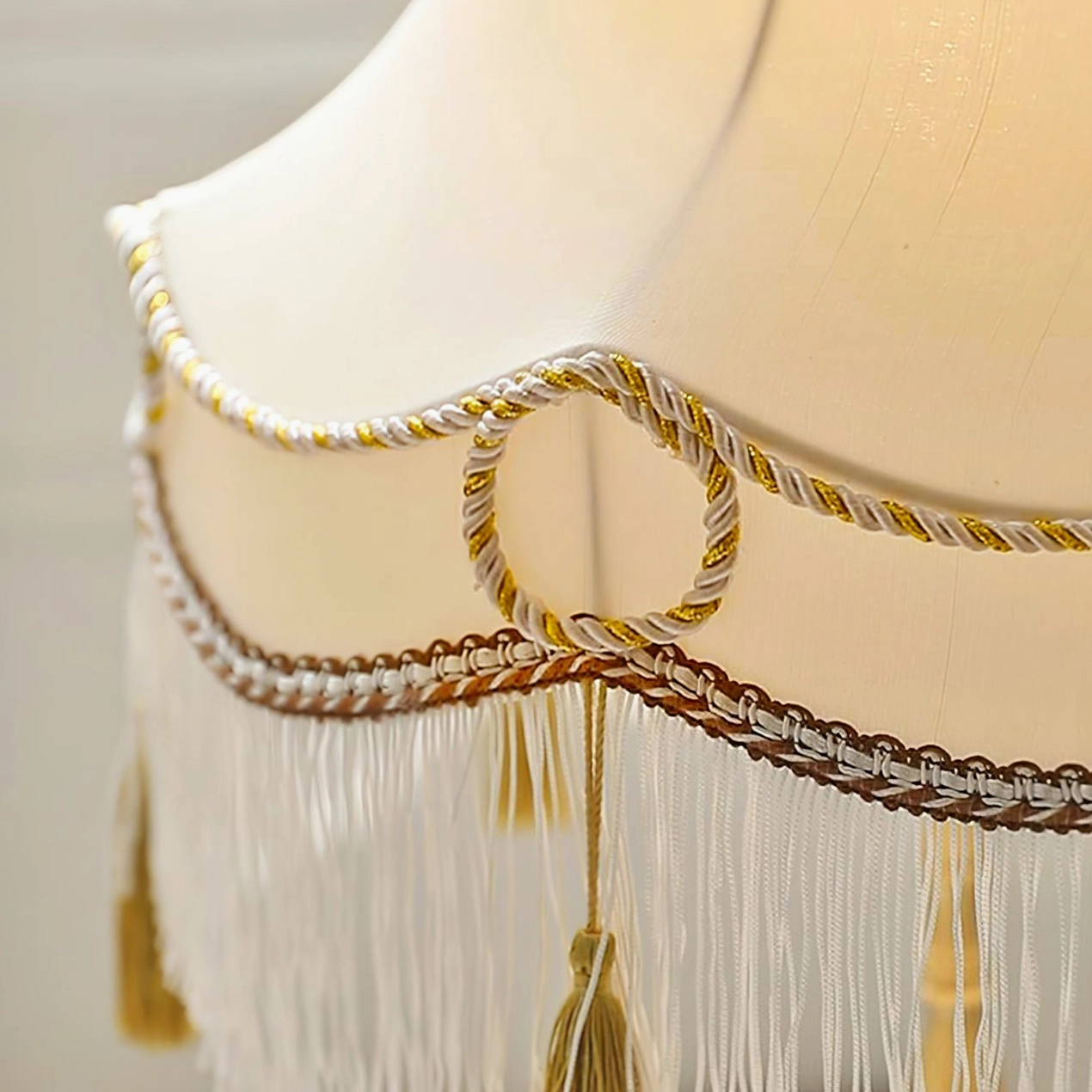 Side angle view of fabric lampshade showing beige and gold-brown braided trim with hanging tassels