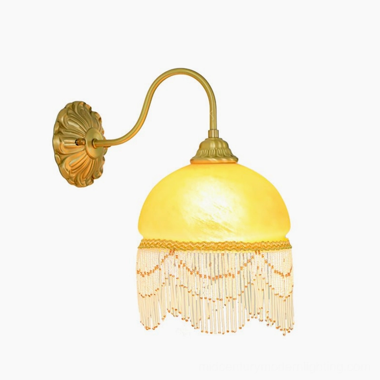 Vili brass wall lamp fully illuminated low front angle, dome shade with gold trim and fringe tassels glowing softly