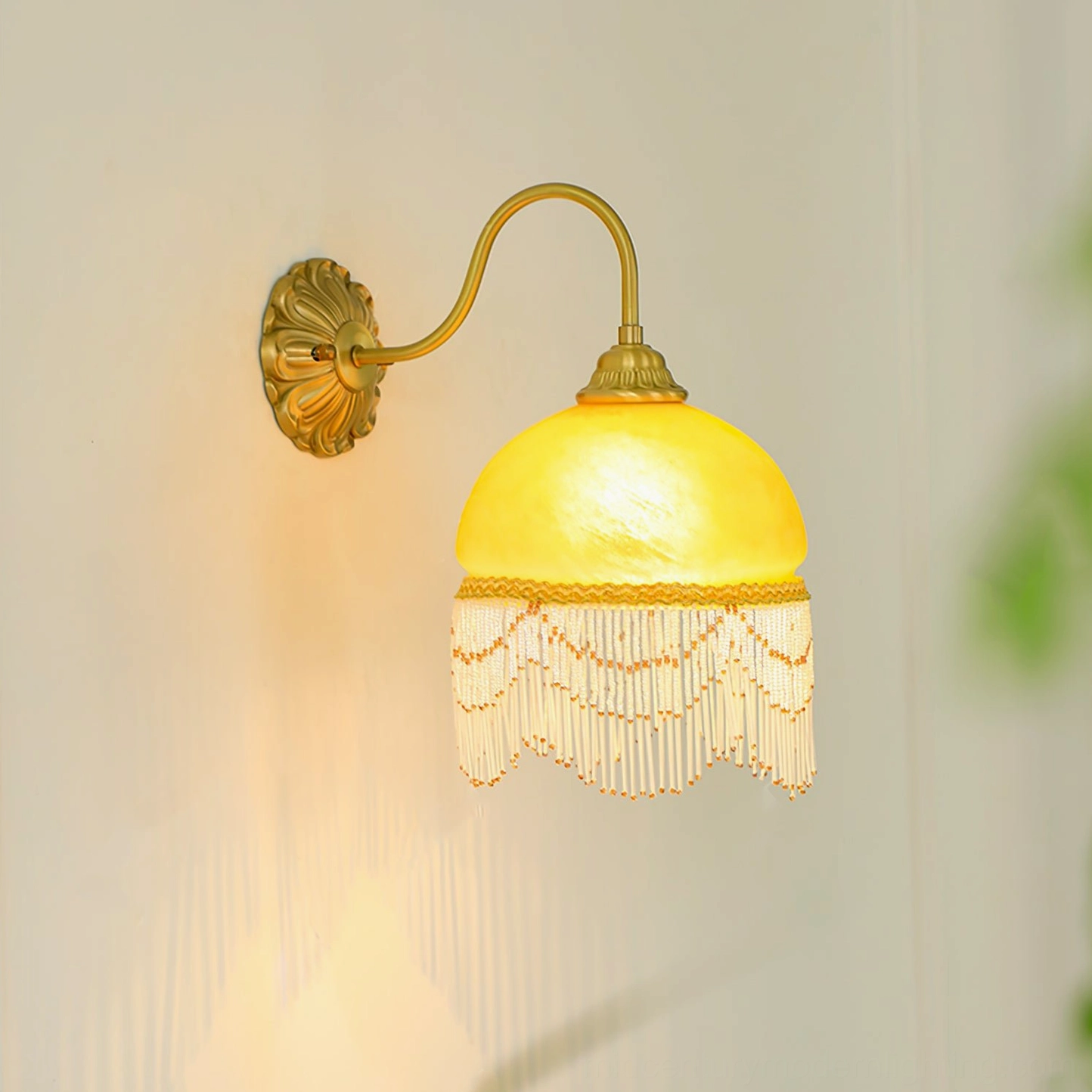 Illuminated Vili wall lamp mounted on green foliage wall, warm glowing light showcasing carved brass base and ambient design