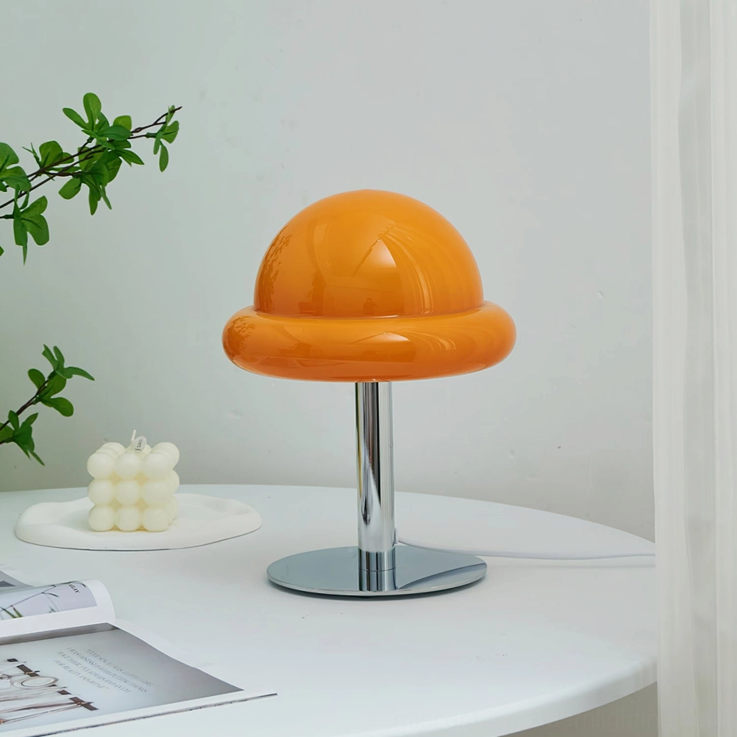 Orange retro table lamp displayed on side view with green plants, candle, magazine, and yellow cup accessories