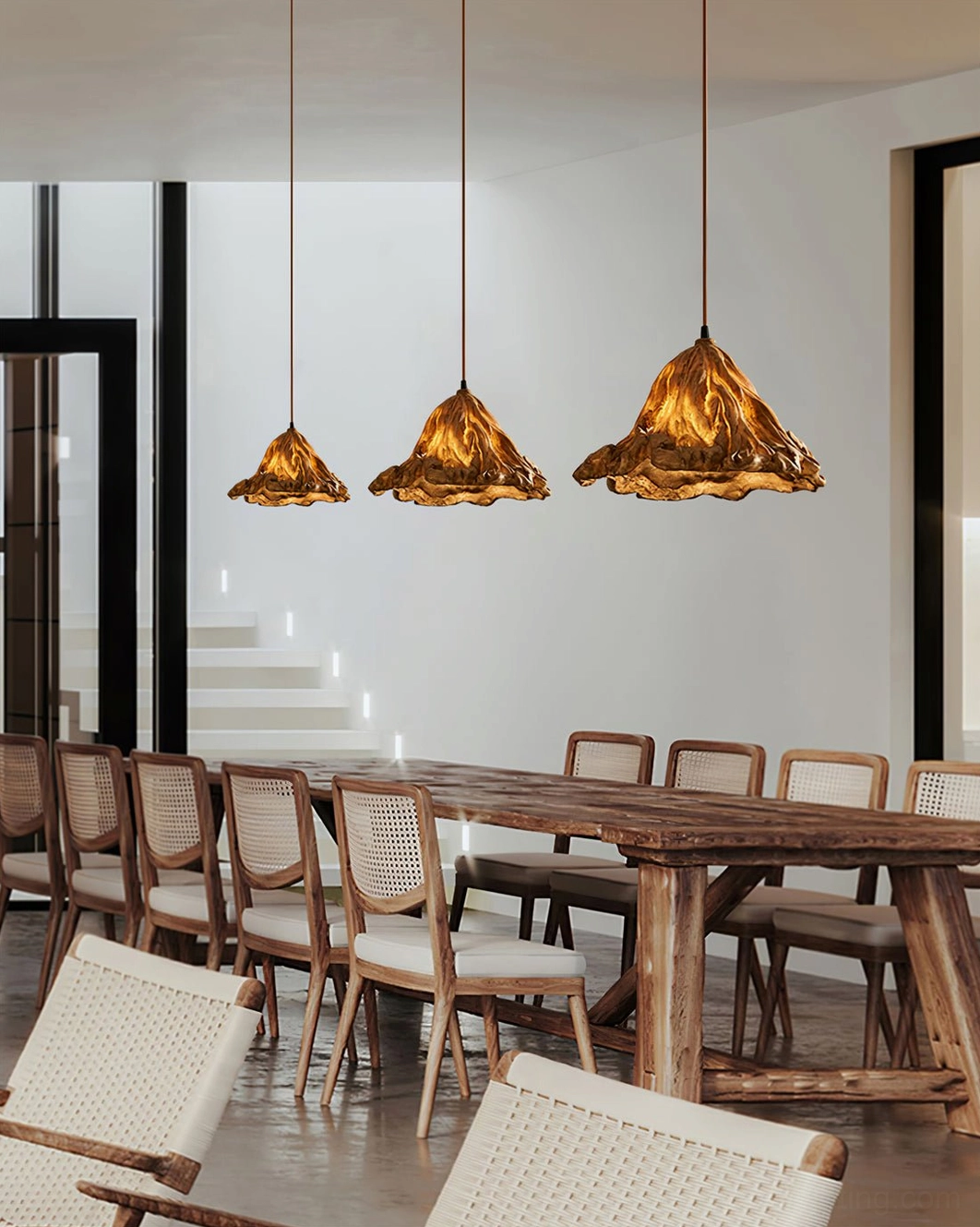 Three brown Volcano pendant lamps over long wooden dining table in minimalist modern dining space with woven chairs