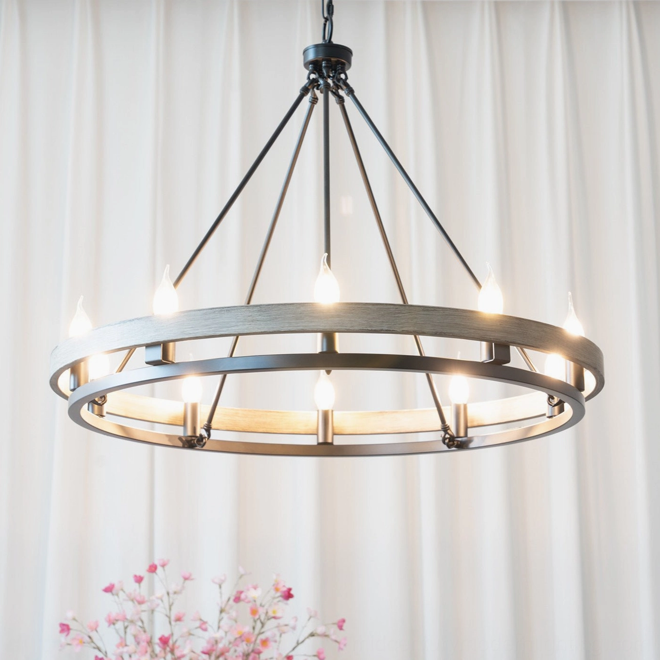 Illuminated wagon wheel chandelier with 10 candle bulbs glowing warm light in rustic interior with dual ring design and sheer curtains