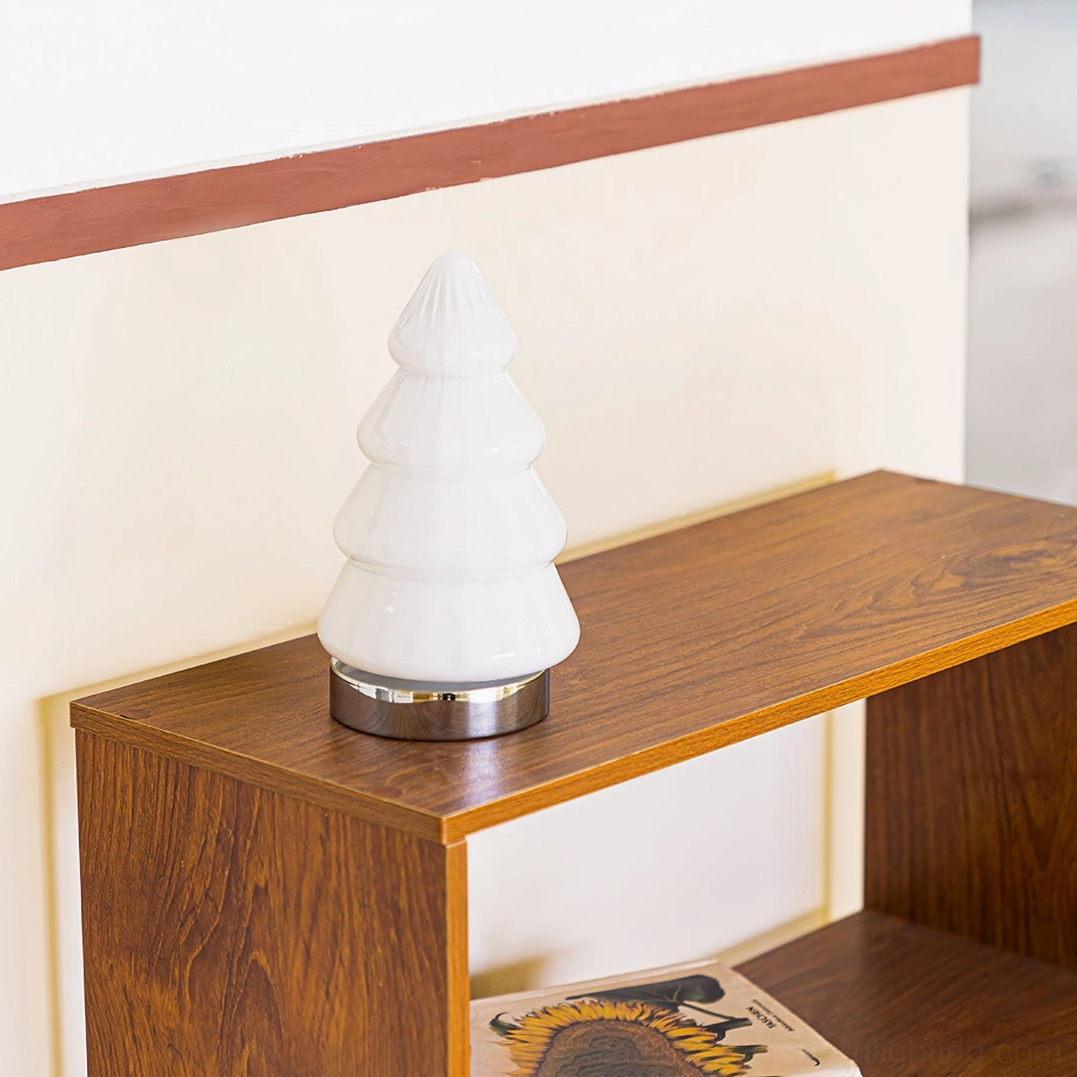 Three-quarter view of white tree table lamp on wooden shelf with warm interior wall trim and framed artwork