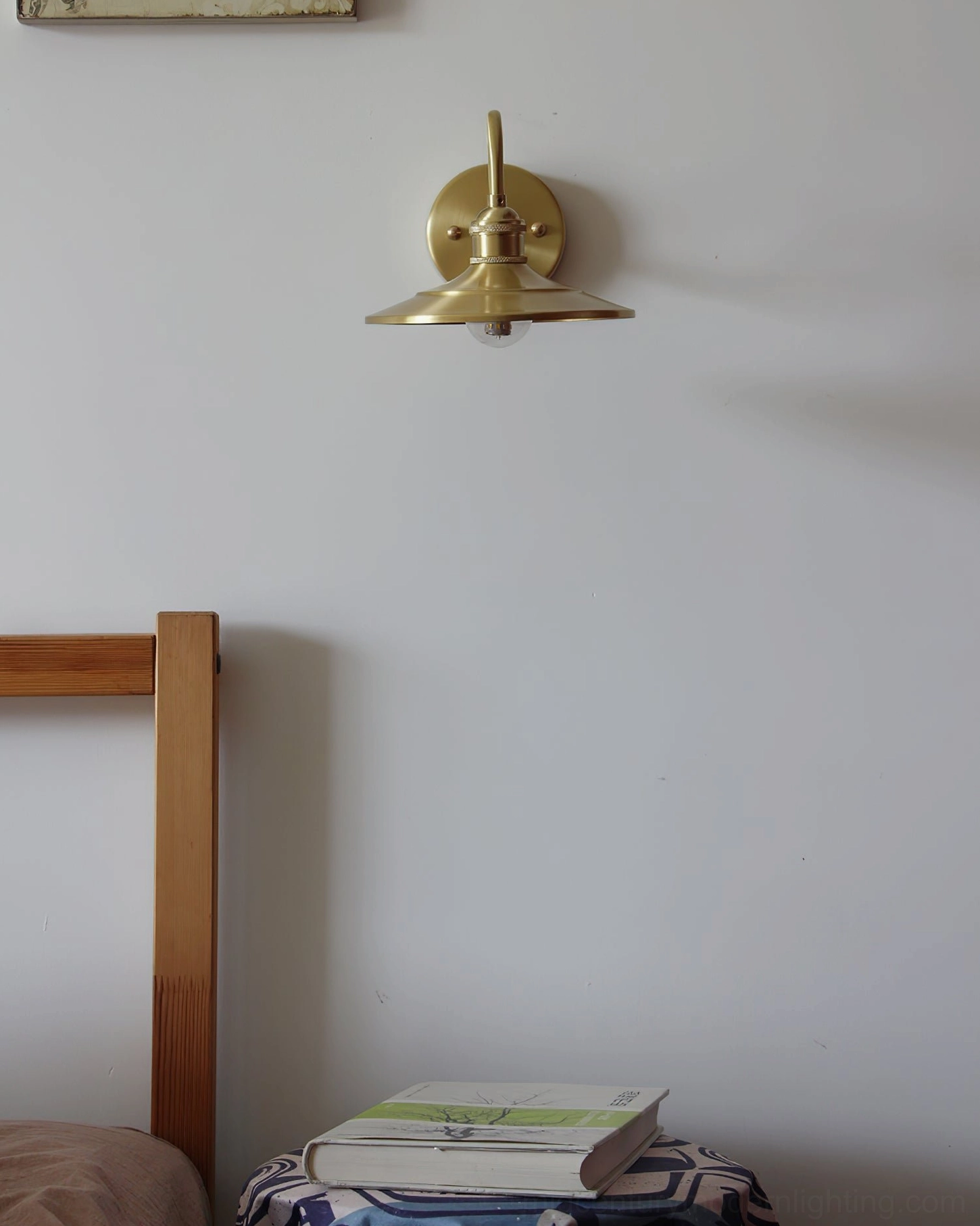 Compact Whizzer brass wall light in minimalist bedroom interior with stacked reading books, neutral wall art, and ambient lighting
