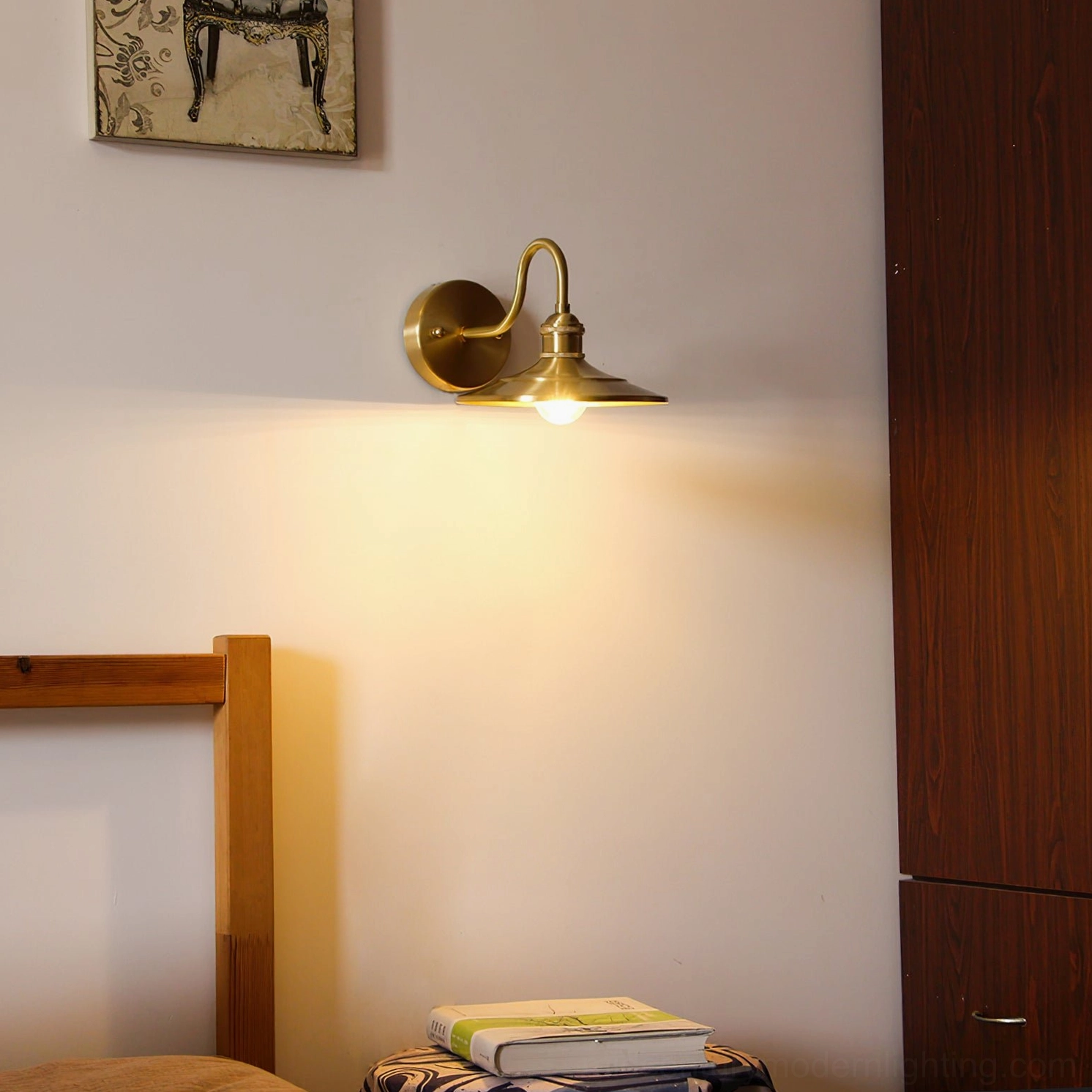 Whizzer brass wall lamp in bedroom corner with warm light, wooden bed frame, and stacked bedside books creating cozy reading nook