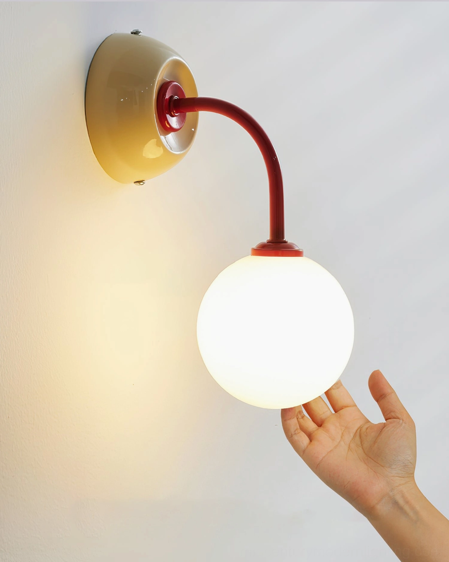 Close-up side view of Zesta wall sconce showing glossy warm-toned metal finish and spherical glass diffuser, hand model demonstrating fixture details